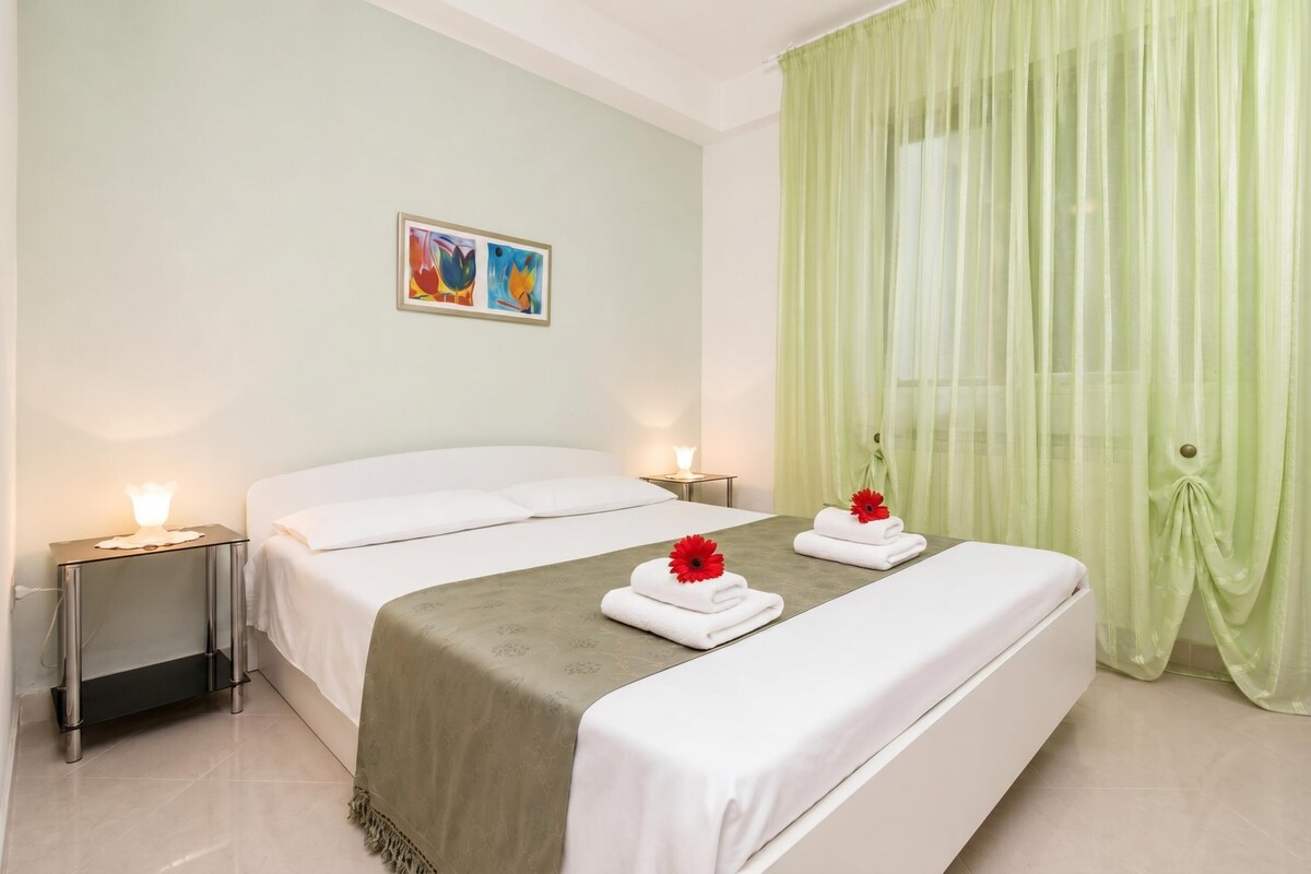 A hotel room with two beds, a nightstand with a lamp, a painting on the wall, and green curtains.