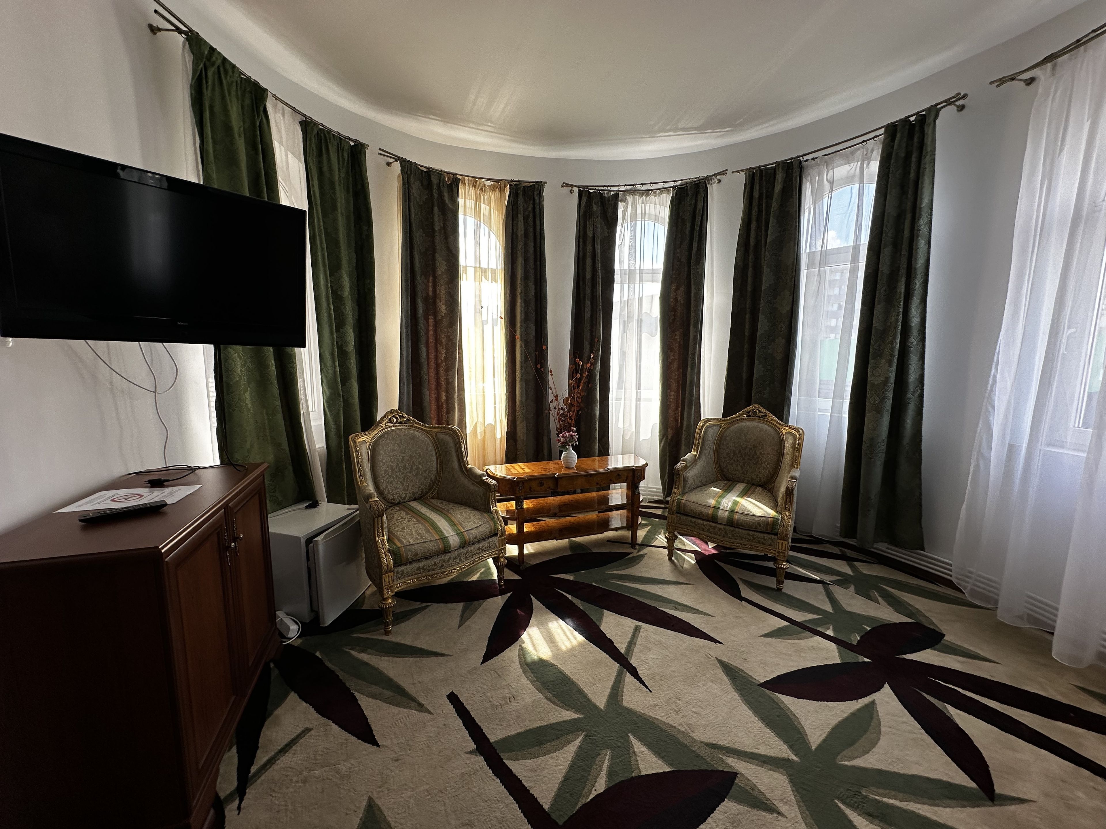Una habitación con una televisión de pantalla plana montada en la pared, dos sillones decorativos, una mesita de madera y una alfombra estampada.