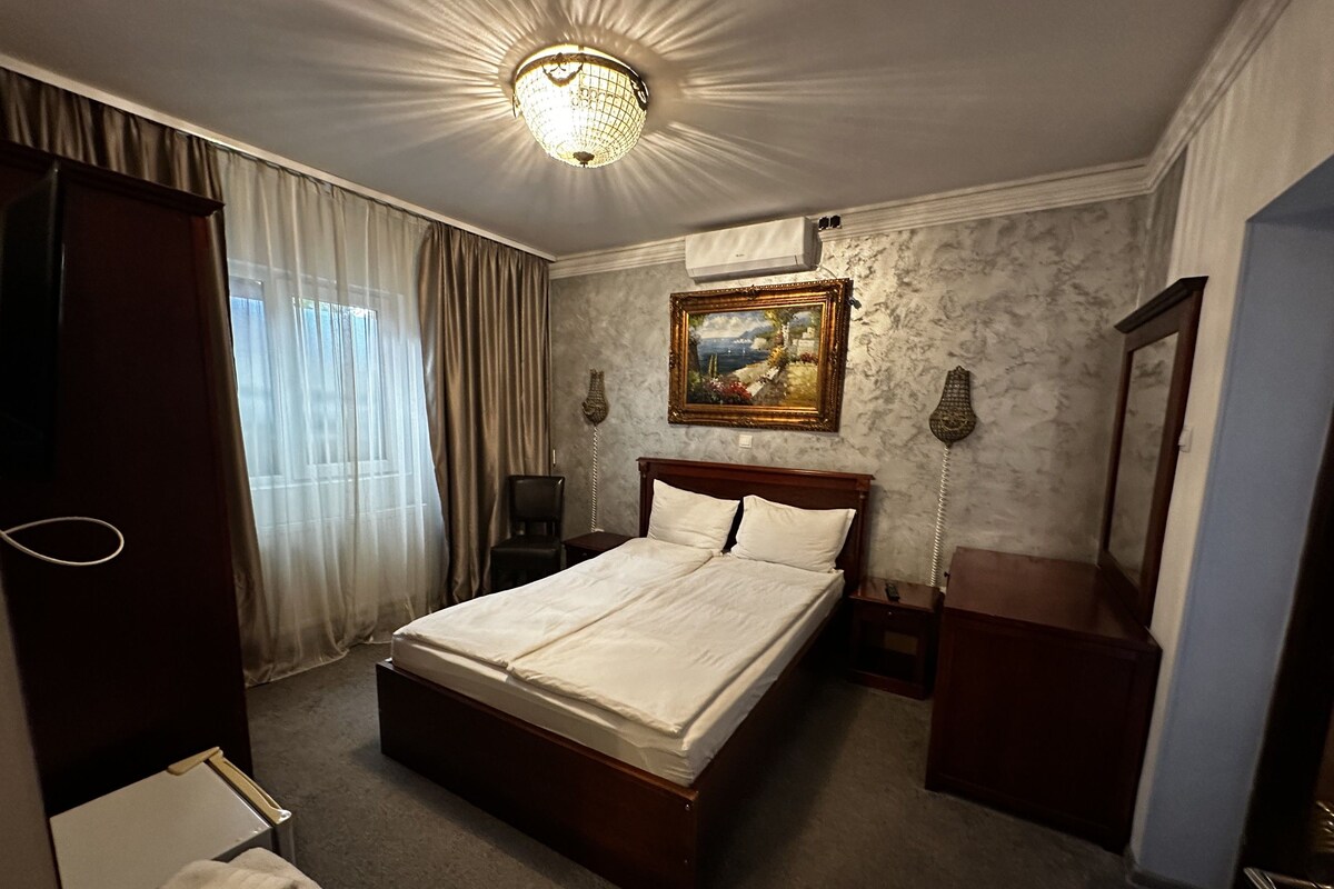 A hotel room with a large bed, a bedside table, a nightstand, a lamp, a painting on the wall, and a ceiling light fixture.