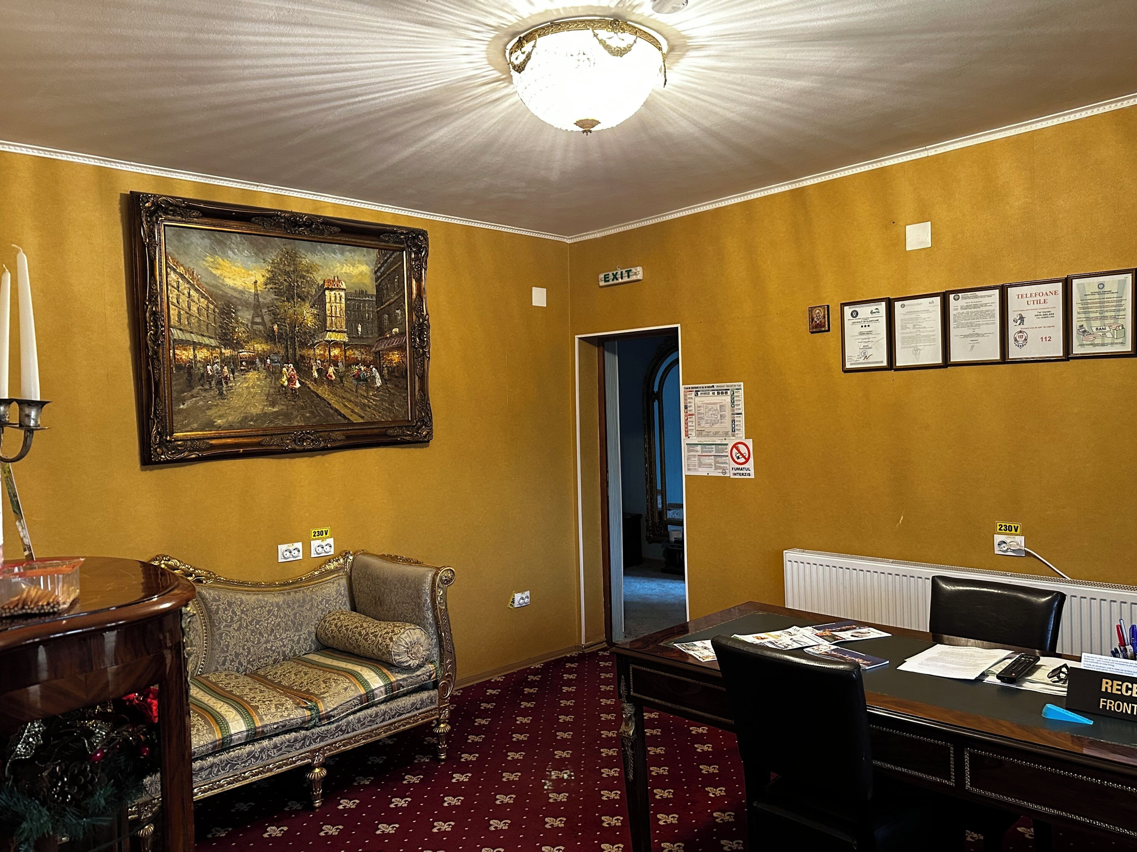 A hotel room with a vintage sofa, a desk with a computer, a framed painting, and a chandelier.