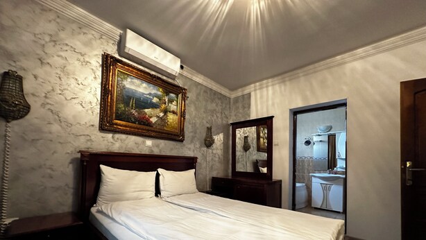 A hotel room with a bed, a painting, a mirror, a bedside table, and a bathroom visible through an open door. - CROWN ROYAL APARTMENTS (Alba Iulia)