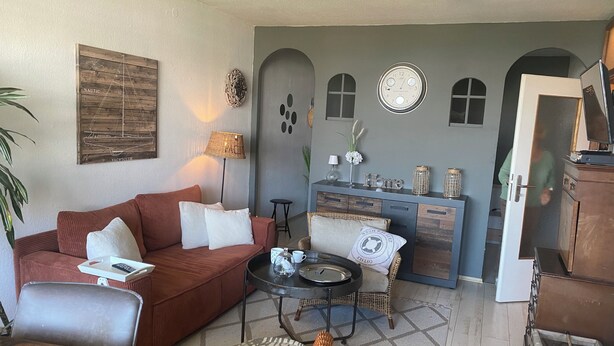 A living room with a red sofa, a coffee table, a wicker chair, a small side table, a lamp, a wall clock, and a cabinet with decorative items. - Beautiful vacation rental in Heiligenhafen with ocean view + balcony through Oct. 2026 (Heiligenhafen)