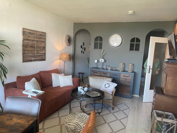 Un salon avec un canapé rouge, une table basse, un fauteuil en osier, une petite table d’appoint, une lampe, une horloge murale et un meuble contenant des objets décoratifs.