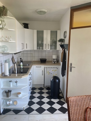 Une cuisine de taille réduite avec des meubles blancs, un sol à carreaux noirs et blancs, une machine à café et une poubelle.