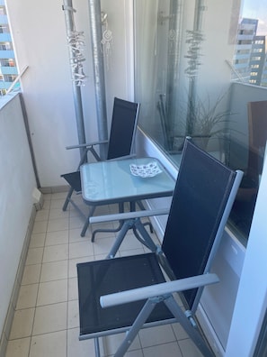 Un coin de pièce avec une table en verre et deux chaises, une plante en pot et une fenêtre donnant sur des immeubles.