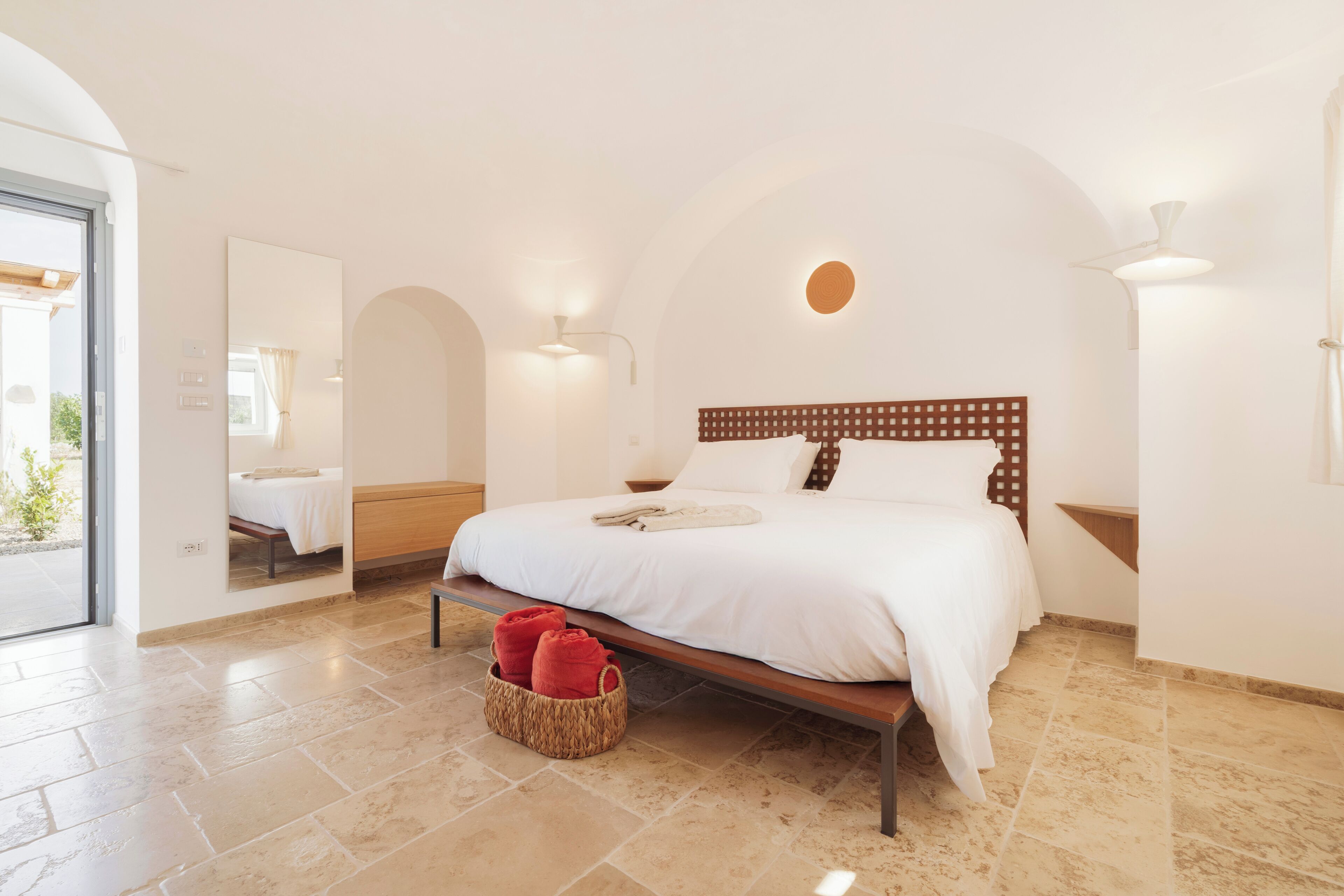 A bedroom with a large bed, a wooden headboard, a bedside table, a wall-mounted light, and a basket with red towels.