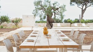 An outdoor dining area with a long table, white chairs, and a large tree in the background. - Trulli Vigna Lamafico Suites & Pool (Polignano a Mare)