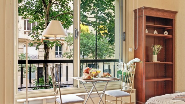 Ein Zimmer mit Balkon, einem Esstisch mit Stühlen, einer Lampe und einem Holzregal.