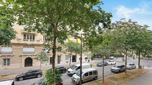 Straßenansicht mit parkenden Autos, Bäumen und einem Gebäude mit Balkonen.