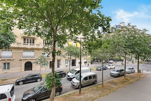 Straßenansicht mit parkenden Autos, Bäumen und einem Gebäude mit Balkonen.