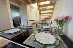 Ein Esstisch ist gedeckt mit Tellern, Gläsern und Besteck, dazu steht eine Vase mit Blumen, und im Hintergrund sieht man eine Holztreppe.