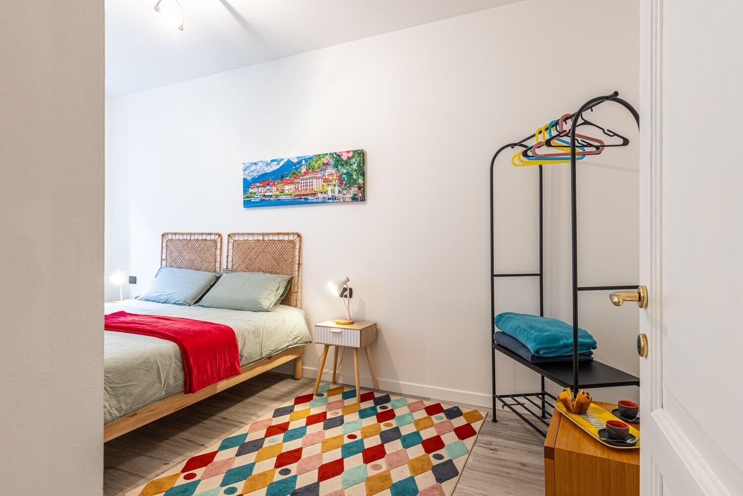 A bedroom with a bed, a colorful rug, a painting, a metal shelf with clothes, and a small table with a tray.