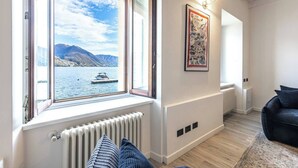 Ein Zimmer mit großem Fenster und Seeblick, einer Heizung, einer schwarzen Couch und einem gerahmten Bild an der Wand.