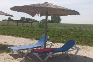 Two beach chairs under a thatched umbrella on a sandy beach with a fence and greenery in the background. - Holiday home in Burhave with swimming pool (Burhave)