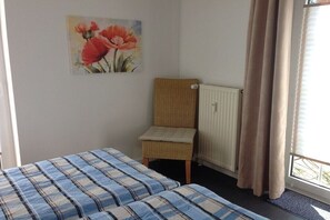 A room with two beds, a wicker chair, a radiator, and a window with curtains. - Holiday apartment with 1 bedroom (Cuxhaven-Döse)