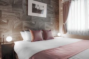 Una habitación de hotel moderna con una cama grande, una mesita de noche con lámpara, un cuadro enmarcado en la pared y una ventana con cortinas.