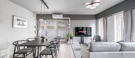 A modern living room with a sofa, a dining table with chairs, a TV, and a large window with curtains.