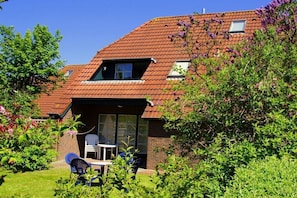 Ein Haus mit geneigtem Dach, einer kleinen angeschlossenen Garage und einem Garten mit Sitzgelegenheiten.