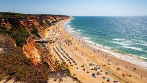 A coastal beach with a long stretch of beach umbrellas and sun loungers, a clear sky, and a rocky cliffside. - Central Albufeira Stay Balcony AC (Albufeira)