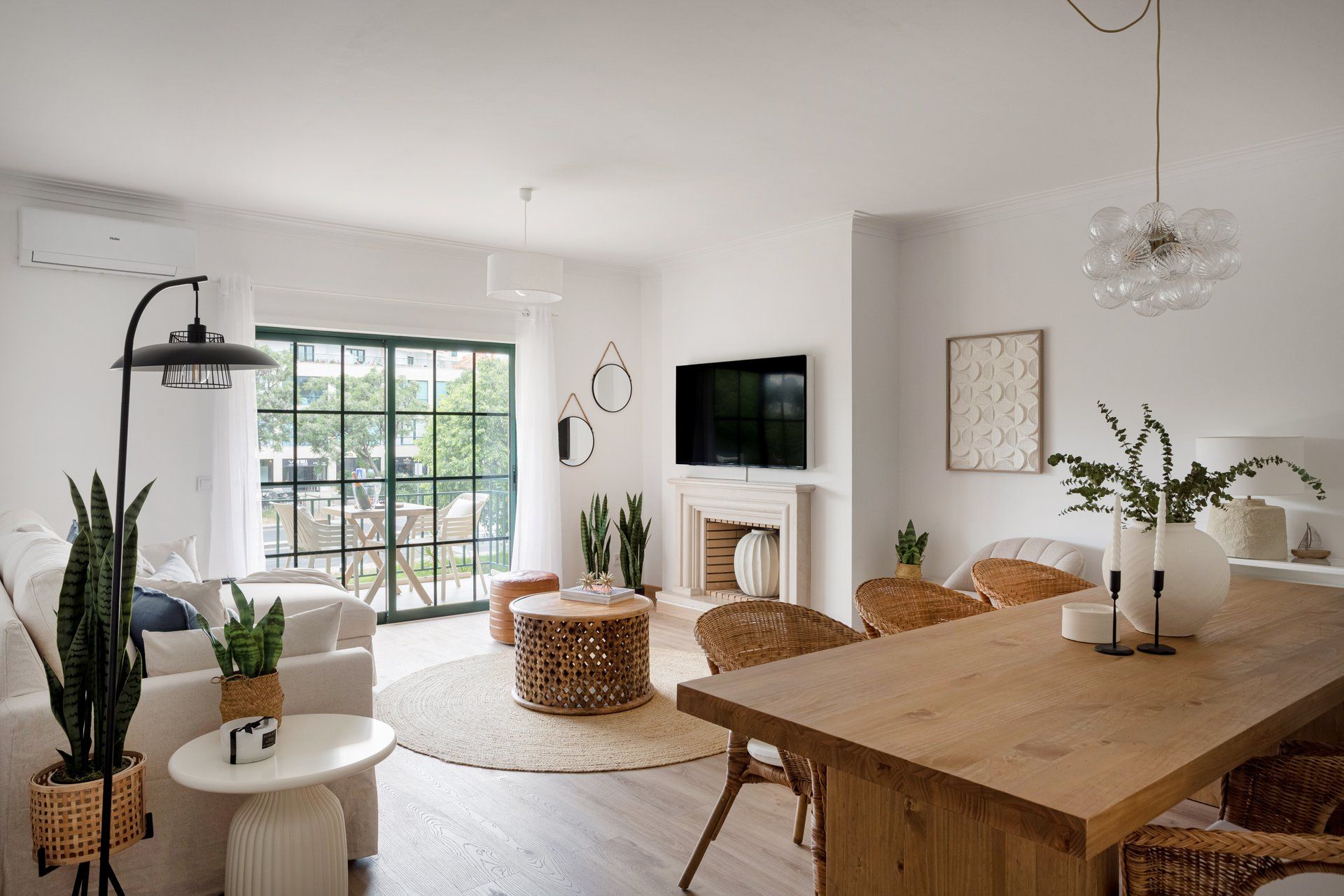 A modern living room with a fireplace, a dining table, wicker furniture, and a large window.