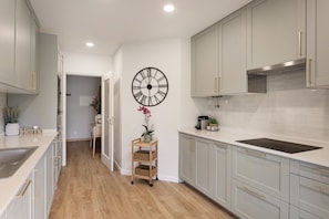 A modern kitchen with light grey cabinets, a black clock, and a small wooden cart with baskets. - Carcavelos Terrace Beach Apartment (Cascais)