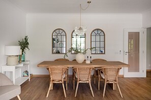 A dining room with a wooden table, wicker chairs, a chandelier, and two arched windows with mirrors. - Carcavelos Terrace Beach Apartment (Cascais)
