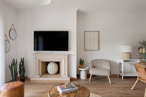 A living room with a fireplace, a flat-screen TV, a round coffee table with magazines, a grey armchair, and a wicker chair. - Carcavelos Terrace Beach Apartment (Cascais)