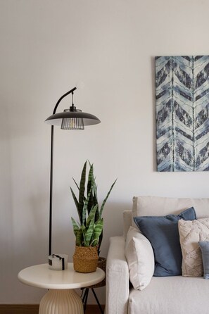 Una sala moderna con una mesa redonda blanca, una lámpara de pie, una planta en maceta, un sofá con cojines y un cuadro abstracto colgado en la pared.