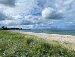 Een strand met een grasveldje in de voorgrond, een metalen hek langs de kust, en een bewolkte lucht.