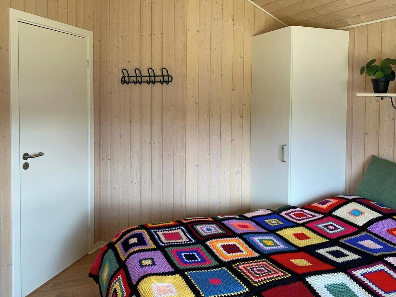 A room with a bed, a door, a wall-mounted hook, and a shelf with a plant.