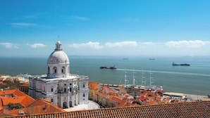 Paisagem costeira com um edifício branco de cúpula imponente, um porto com navios e um céu azul claro.