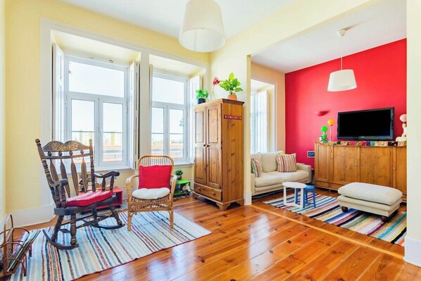 A living room with a wooden floor, a rocking chair, a sofa, a television, and a red wall. - Stylish Lisbon Flat in Graça With Terrace and View (Lisbon)