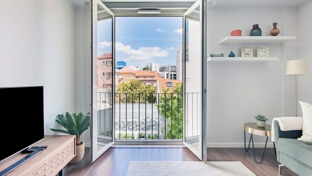 Ein modernes Wohnzimmer mit Fernseher, einer Pflanze, einem Balkon mit Blick auf Gebäude und einem kleinen Beistelltisch.
