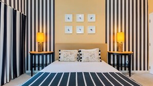 Hotellihuone, jossa on raidallinen seinä, raidallinen päiväpeite sängyllä, kaksi yöpöytää valaisimineen ja seinälle kiinnitetty kello.