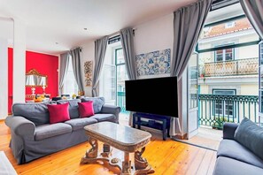 Ein Wohnzimmer mit grauem Sofa, einem Holztisch, einem Flachbildfernseher und einem Balkon mit Geländer.