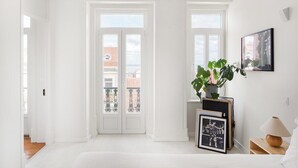 Een witte kamer met een bed, een potplant, een ingelijst plaatje en een lamp op een bijzettafeltje.