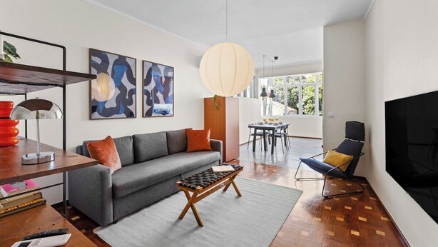Ein modernes Wohnzimmer mit Sofa, Couchtisch und Fernseher. An den Wänden befinden sich abstrakte Gemälde und im Hintergrund ein großes Fenster.