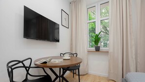 Een kamer met een aan de muur bevestigde televisie, een ronde tafel met stoelen, een raam met gordijnen en een bed.