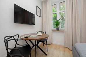 Ein Zimmer mit Fernseher an der Wand, einem runden Tisch mit Stühlen, einem Fenster mit Vorhängen und einem Bett.