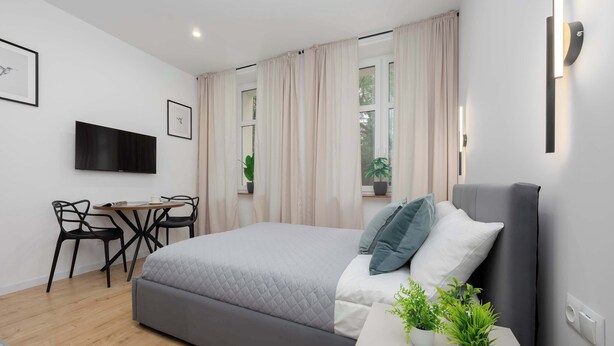 Een moderne slaapkamer met een bed, nachtkastje, potplanten, een bureau met stoelen en een televisie aan de muur.