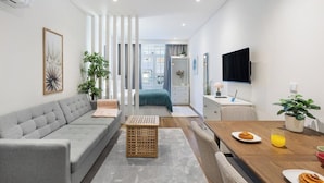 Ein modernes Wohnzimmer mit Sofa, Esstisch, Geschirr und einem Glas, einem Fernseher und Blick ins Schlafzimmer.