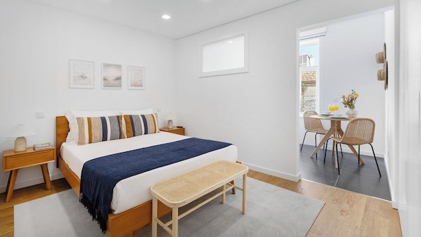 A bedroom with a bed, bedside tables, a bench, a dining area with chairs, and a window. - Matosinhos Coastal Breeze Studio (Matosinhos)