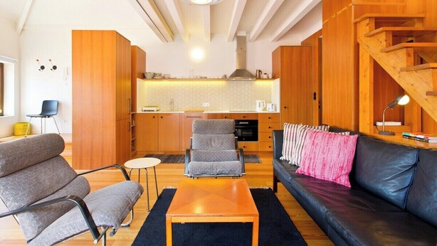 Een moderne woonkamer met een bank, fauteuil en salontafel, met een keuken op de achtergrond en een houten trap.