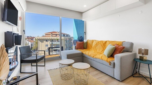 A modern living room with a grey sofa, a flat-screen TV, a balcony with a view of buildings, and a large window. - Blue Breeze Espinho Pool View, Gym Balcony (Espinho)
