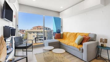 A modern living room with a grey sofa, a flat-screen TV, a balcony with a view of buildings, and a large window.