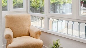 A room with a large window, a beige armchair, a small round table with a plant, and a woven blind. - Estoril Beach Your Family Escape (Cascais)