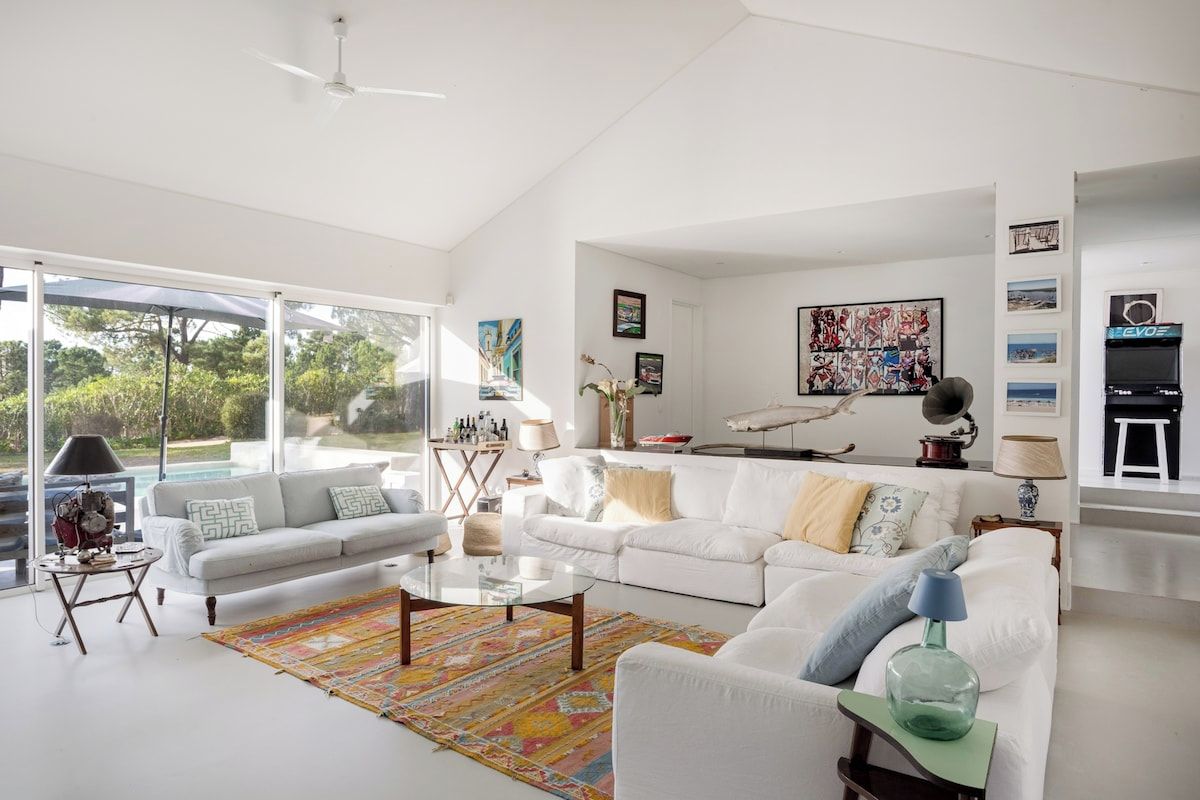 A spacious living room with a white sofa, a coffee table, and a colorful rug. There is a large window providing a view of the outdoors.