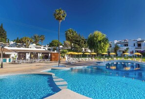 A swimming pool with lounge chairs and umbrellas, surrounded by buildings and palm trees. - Clube Albufeira - 2BR Pool, Kids Club Family Fun (Albufeira)