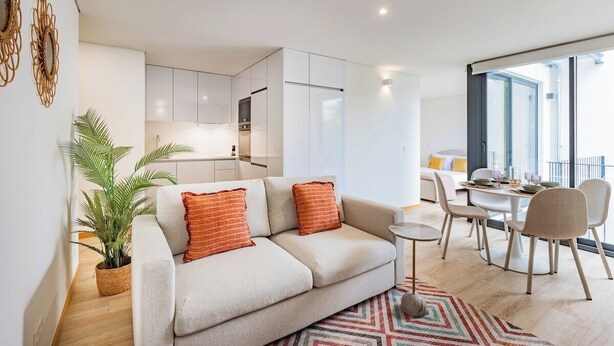 A modern living space with a sofa, dining table, chairs, a kitchen area, and a large window. - Duque de Saldanha Residences, Unit F (Porto)