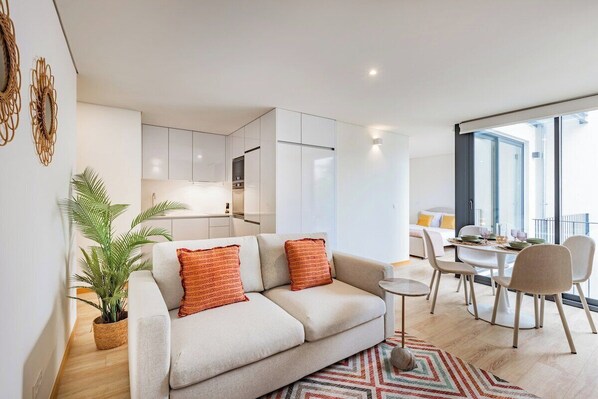 Ein modernes Wohnzimmer mit Sofa, Esstisch, Stühlen, Küchenzeile und großem Fenster.